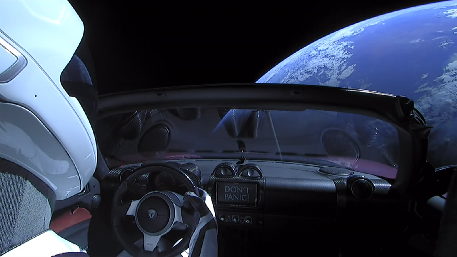 View over Starman's shoulder showing the steering wheel and a DON'T PANIC sign on the dashboard, with Earth beyond