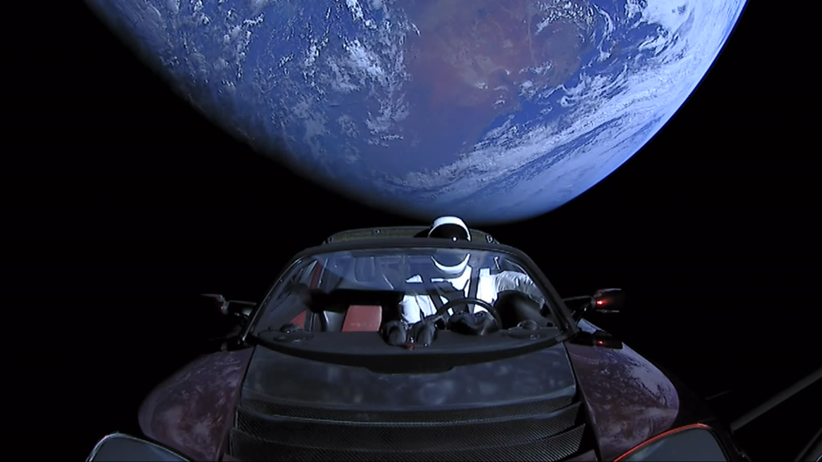 A mannequin in a spacesuit sits in a red Tesla Roadster with Earth filling the sky behind it